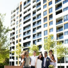 Cute-3D-rendering-of-a-happy-young-couple-standing-in-front-of-a-modern,-affordable-apartment-building-with-green-trees.