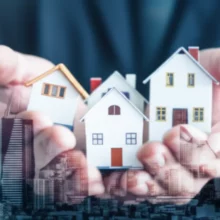 Hands-holding-miniature-houses-above-a-city-background